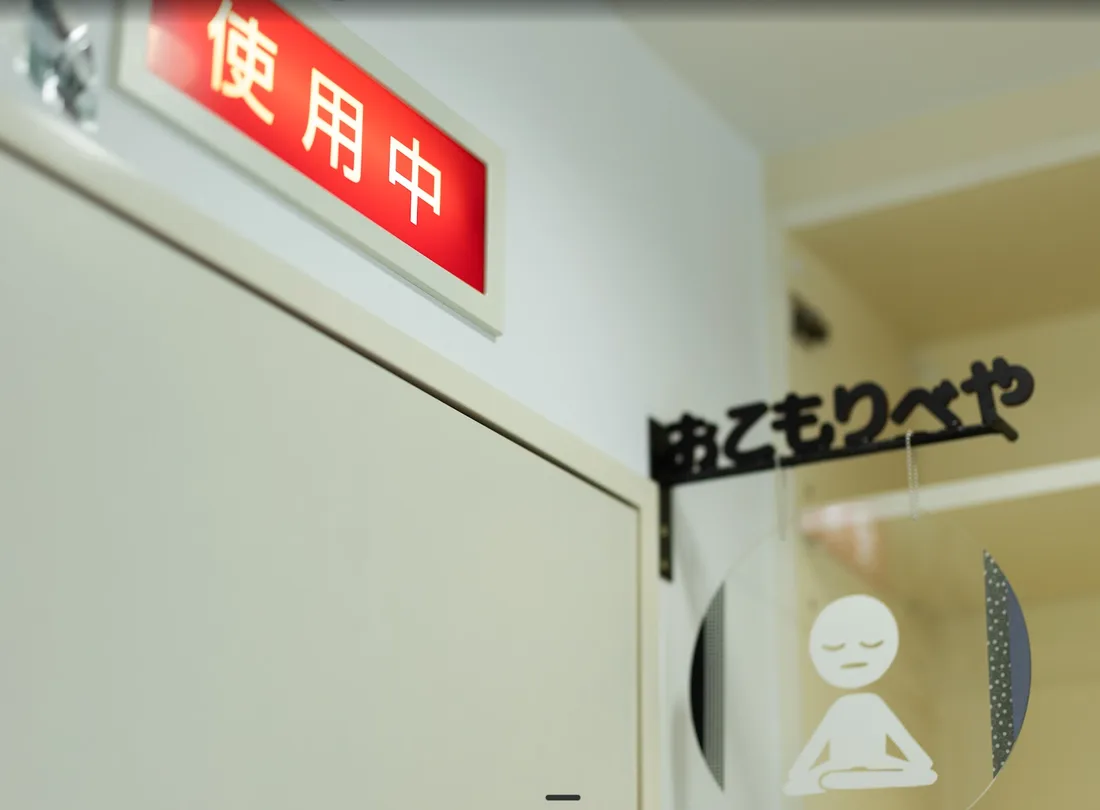 「おこもりべや」と書かれた入口のサイン。上に「使用中」の赤いランプ、ガラス戸には目を閉じて座る人のマーク